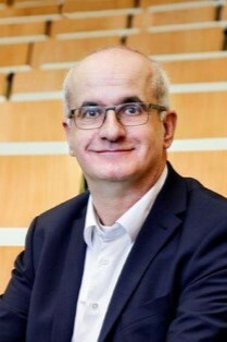 Manfred Bayer mit Brille, Anzug und Hemd, sitzt in einem Hörsaal mit Holzstühlen.